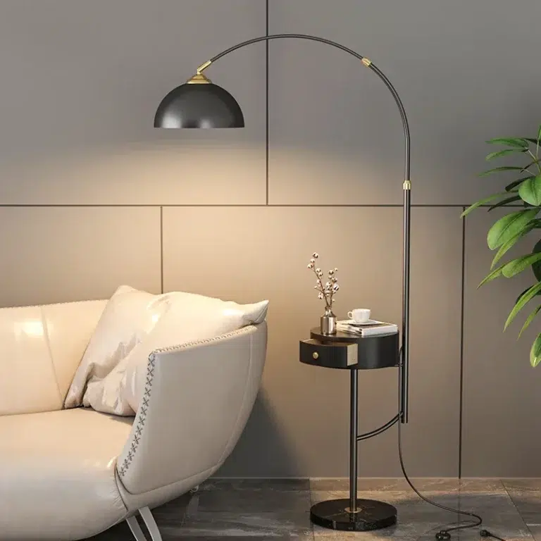 Lampadaire noir avec socle rond surmonté un support rond avec tiroir puis un arc avec abat-jour noir, ampoule allumée. Un canapé blanc en similicuir à gauche, une plante à droite. Un livre, une tasse blanche et une fleur de coton sur le support du lampadaire.