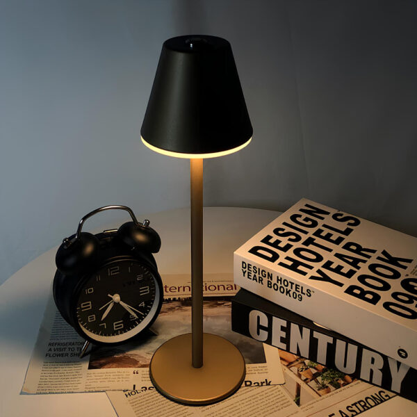 Lampe champignon noir avec long pied fin et socle rond. Posée sur une table ronde blanche. A sa gauche, un réveil vintage noir, à sa droite, deux livres l'un sur l'autre. La table est contre un mur gris.
