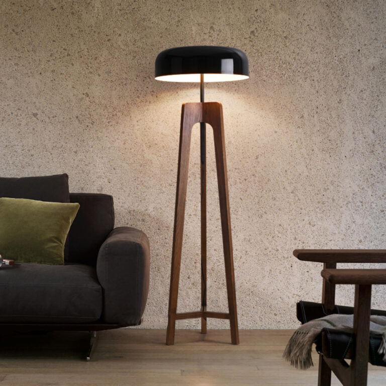 Lampadaire champignon avec chapeau noir en métal et pied en bois. Il est allumé et posé à droite d'un canapé gris, contre un mur beige. Le devant d'un fauteuil en bois est visible devant à droite.