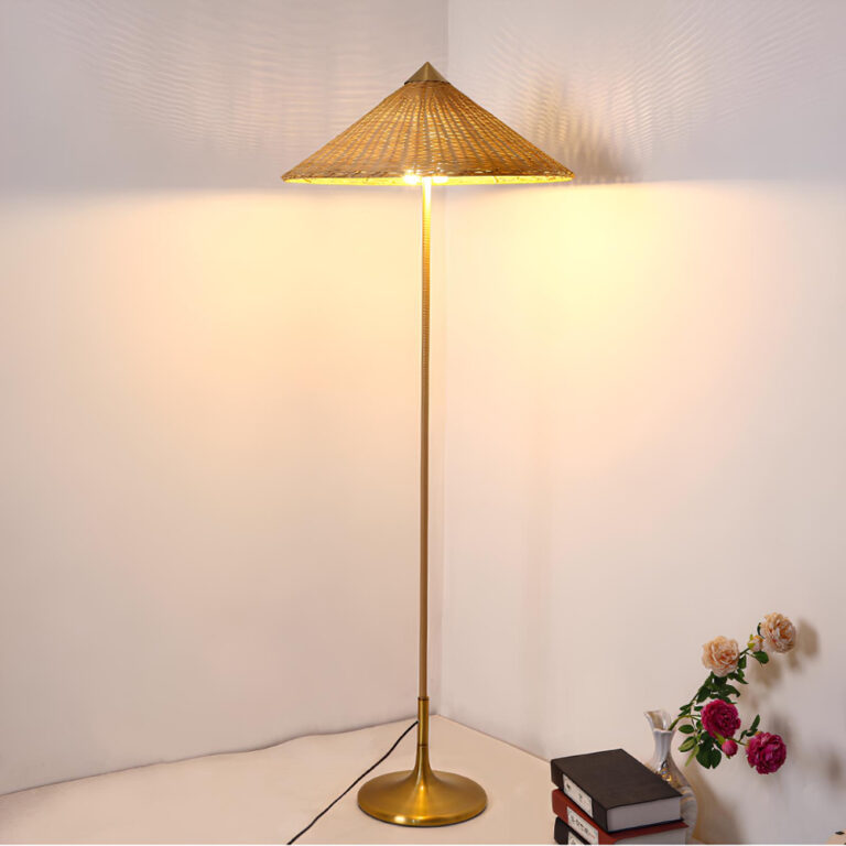 Lampadaire champignon avec son chapeau pointu en rotin. Son socle au sol est en métal doré. Son pied est droit et vertical. Il est allumé, placé dans un angle de murs. A droite au sol, un vase blanc avec des fleurs roses et des livres empilés.
