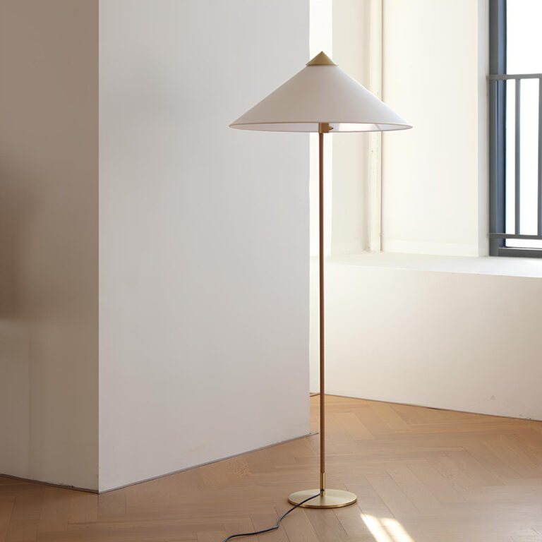 Lampadaire avec socle rond doré, un pied fin doré et un abat-jour pointu en tissu blanc avec la pointe dorée. Dans une pièce vide, au sol marron clair et aux murs blancs.