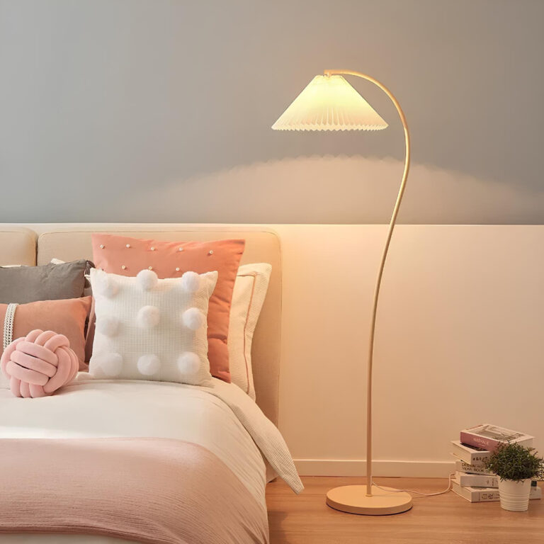 Lampadaire champignon avec socle rond beige, pied arrondi avec abar-jour plissé en forme de chapeau blanc. L'ampoule est allumée. Le lampadaire se trouve dans une chambre, à droite d'un lit avec des coussins de plusieurs couleurs et un drp blanc. Le mur est séparé en 2 : la partie basse est blanche et la partie haute est grise.