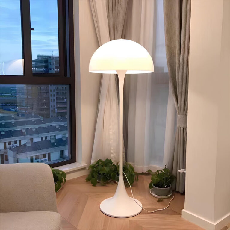 Lampadaire champignon blanc avec lumière allumée. Il se trouve devant des grandes fenêtres dont une avec un voilage blanc. Le sol ressemble à du parquet flottant beige.