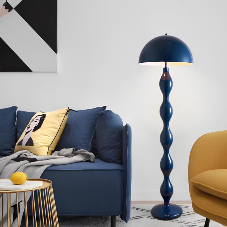 Lampadaire champignon en métal bleu foncé avec un pied aux courbes ondulées. A sa gauche, un canapé bleu foncé avec un coussin jaune et devant lui une table basse style rotin. A droite du lampadaire, un fauteuil jaune moutarde. Le mur derrière est blanc avec un cadre au-dessus du canapé bleu.