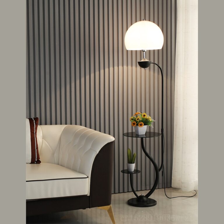 Lampadaire en métal noir fin avec 2 supports, un petit en bas et un plus grand au-dessus, faisant office de tables d'appoint, surmontés d'une tige noire avec une demi-sphère lumineuse en haut. Le lampadaire est à droite d'un canapé marron et blanc. Un mur gris derrière, le sol est du carrelage blanc.