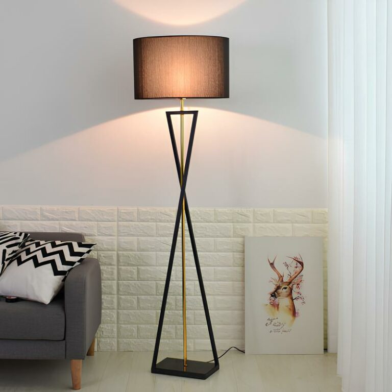 lampadaire champignon avec abat-jour rectangulaire noir, l'ampoule est allumée et sa lumière est visible à travers l'abat-jour. La structure est en métal croisé noir avec une tige dorée au centre. A droite, au sol, un cadre, à gauche, un canapé gris avec des coussins blancs et noirs. Le mur est blanc.
