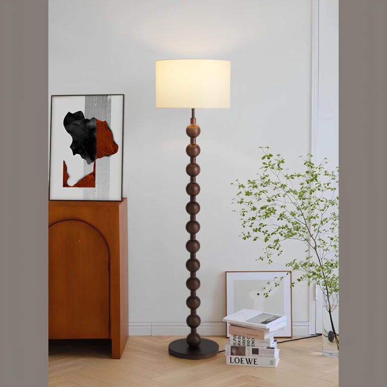 Lampadaire champignon avec socle et pied en bois naturel. Le pied est formé de boules empilées. Son abat-jour blanc rectangulaire diffuse une lumière blanc chaud. Le lampadaire se trouve à droite d'un meuble en bois sur lequel se trouve un cadre abstrait rouge, noir et blanc. A droite du lampadaire, une pile de livres au sol, un cadre carré contre le mur blanc et une plante verte fine.