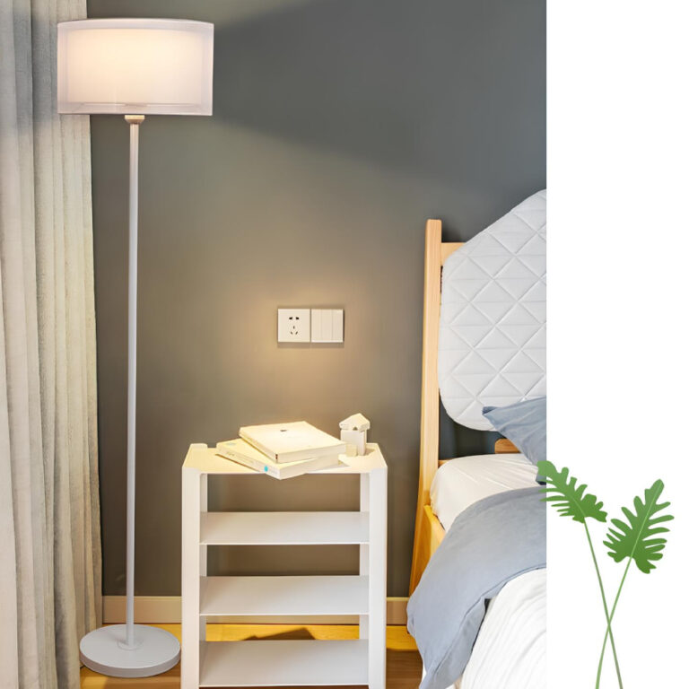 Lampadaire blanc avec abat-jour rectangulaire blanc en tissu. Il est allumé et est à côté d'une tale de chevet blanche, à sa gauche un rideau gris. A droite de la table de chevet, un lit en bois avec literie bleue et blanche. Le mur derrière est bleu foncé.
