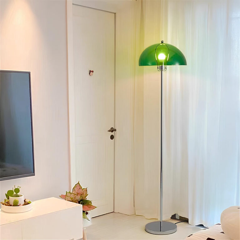Lampadaire champignon avec socle rond et pied de couleur argenté et l'abat-jour en demi-sphère verte avec l'ampoule allumée. Il se trouve dans un salon avec les murs blancs, à sa droite, un rideau blanc, à gauche une porte fermée, encore à gauche un meuble blanc avec un écran.