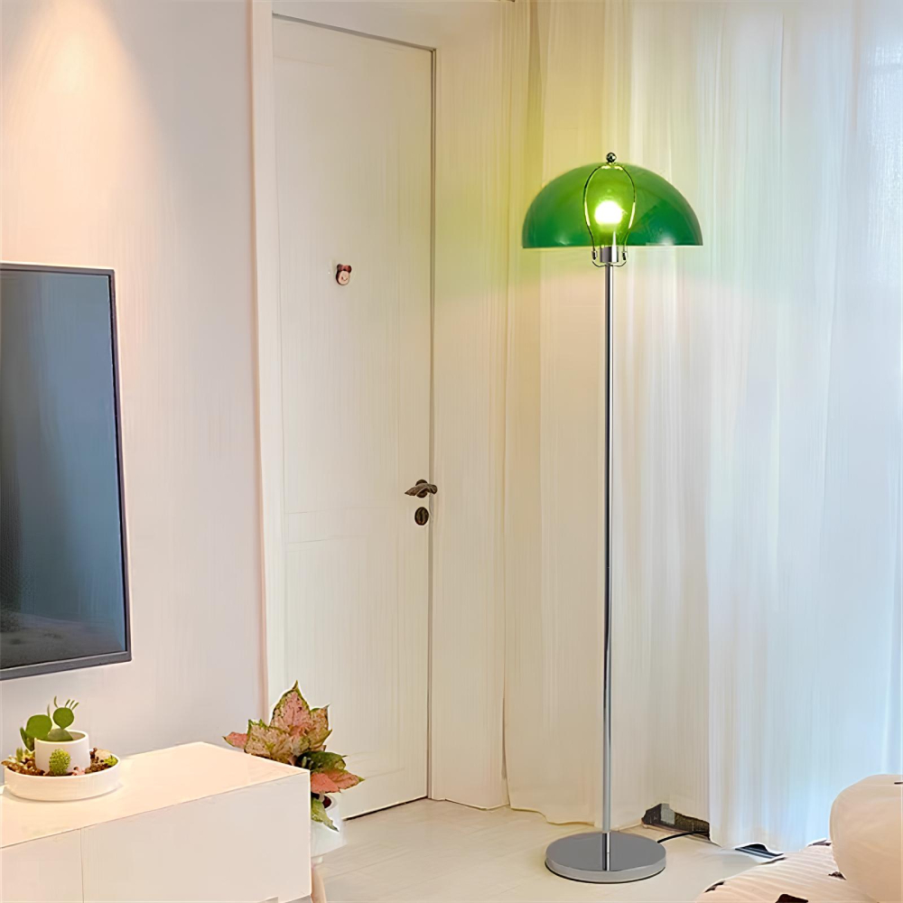 Lampadaire champignon avec socle rond et pied de couleur argenté et l'abat-jour en demi-sphère verte avec l'ampoule allumée. Il se trouve dans un salon avec les murs blancs, à sa droite, un rideau blanc, à gauche une porte fermée, encore à gauche un meuble blanc avec un écran.