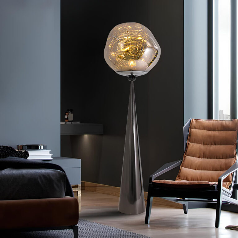 Lampadaire avec un pied noir en forme de cône haut, fin et pointu sur lequel se trouve une sphère lumineuse donnant l'impression d'une boule de lave orangée. Le lampadaire est dans une pièce avec un fauteuil à sa droite marron et orange. Une grande fenêtre sur la droite. Le mur du fond est noir. A gauche, un mur gris avec un meuble bas et le bout d'un lit. Le sol est beige style parquet flottant.