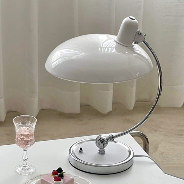 Lampe champignon au design en métal. Le socle rond est blanc avec le contour argenté, la tige est en arc de cercle argentée, l'abat-jour en forme de chapeau de champignon est blanc. Elle est posée sur une table blanche, un verre à pied à sa gauche. Derrière un rideau blanc.