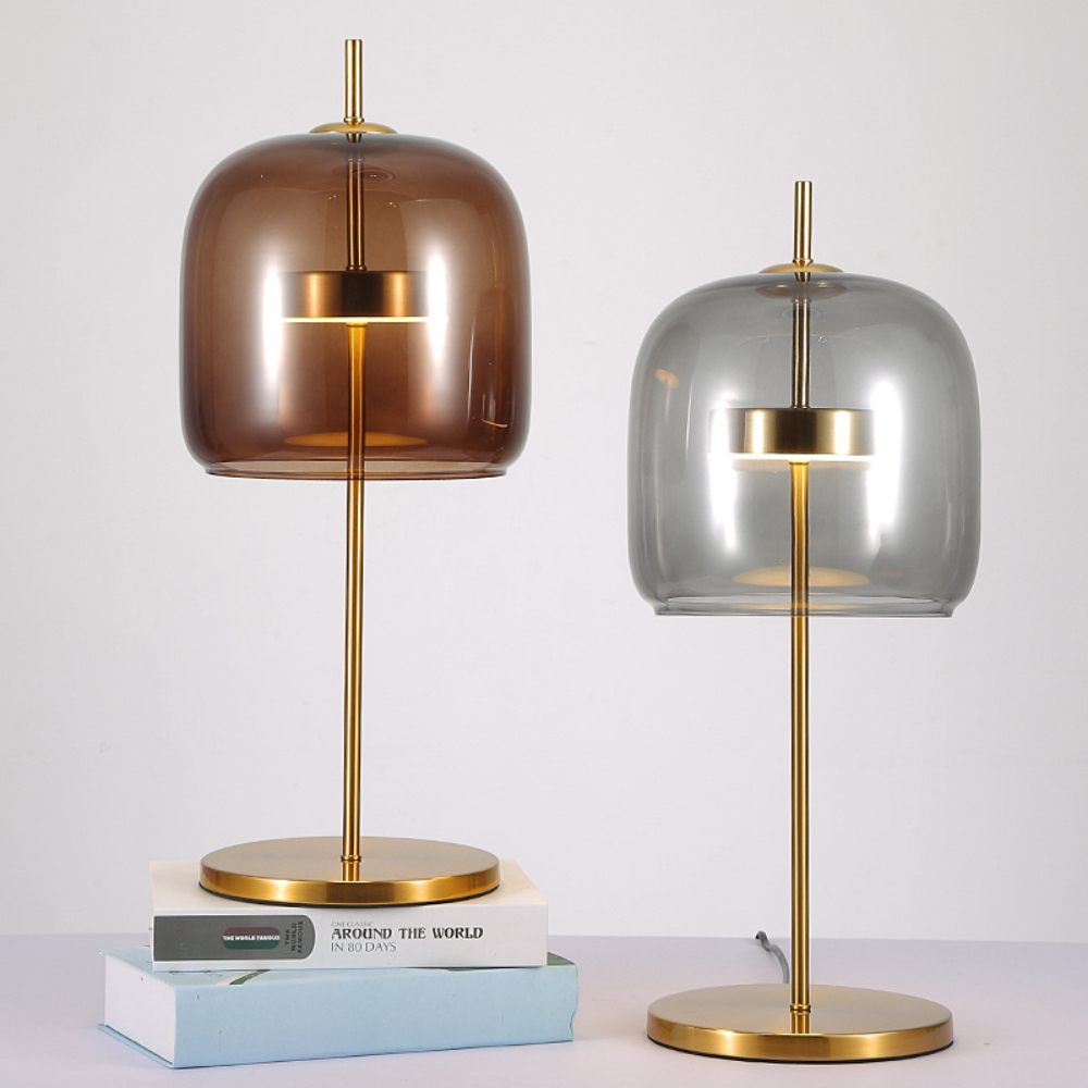 Deux lampes de table champignon avec socle et pied dorés. Celle de gauche à une boule en verre fumé marron et celle de droite en verre fumé gris. La lampe de gauche est surélevée sur des livres. Mur blanc à l'arrière.