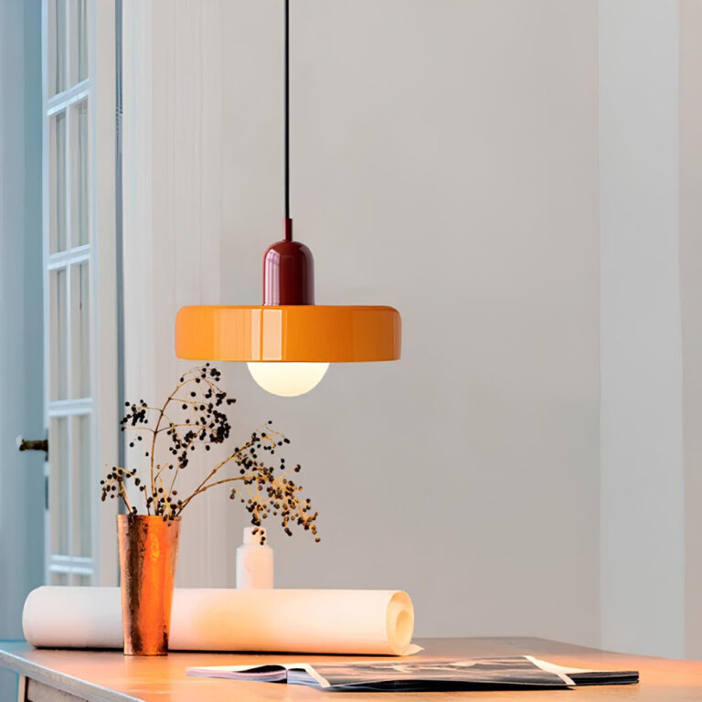 Suspension champignon en verre ressemblant à un disque épais orange, la douille est rouge. L'ampoule est allumée en dessous. Relié par un câble noir. Elle se trouve au-dessus d'une table sur laquelle sont posés un vase un magazine et un papier roulé. Le mur au fond est blanc avec une porte ouverte.