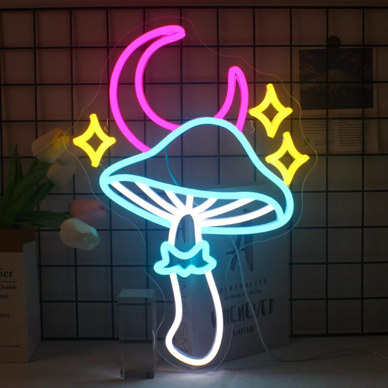 Néon où le champignon est bleu et blanc. Au-dessus 3 étoiles jaunes et un croissant de lune rose. Le néon est allumé et posé contre une grille noire. On voit un livre derrière le néon et des tulipes sur la gauche.