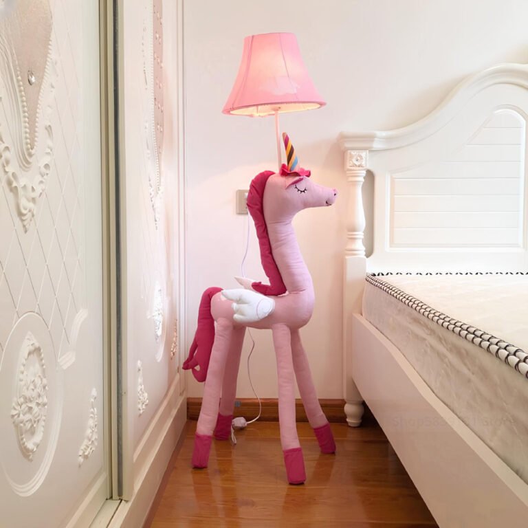 Lampadaire champignon avec licorne rose en tissu. Sur sa tête, on retrouve l'abat-jour rose avec une ampoule allumée. A droite du lampadaire, un lit blanc, à gauche le mur blanc. La tête de la licorne est tournée vers a droite.