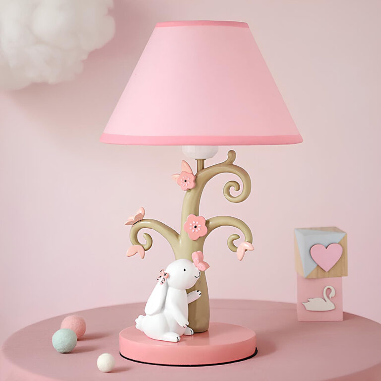 Lampe champignon pour enfant avec socle rond rose sur lequel se trouve une figurine de lapin blanc. Le corps de la lampe ressemble à un tronc avec des branches en spirales. on y trouve des petits papillons roses. L'abat-jour est rose. La lampe est posée sur un table ronde rose. Le mur derrière est rose. A gauche de la lampe, trois petites boules de couleurs pastel et à droite 2 cubes.