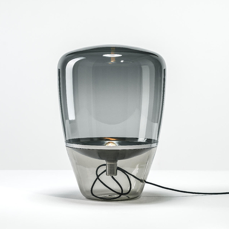 Lampe champignon au design moderne en verre soufflé. La lampe est entièrement en verre fumé gris avec à l'intérieur une coupelle argentée où se trouve l'ampoule. La coupelle est reliée par un fil noir à l'extérieur du verre. Sur fond blanc.