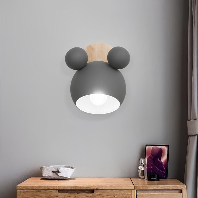 Lampe champignon murale avec socle rond en bois et son abat-jour de forme ronde et de couleur grise rappelle la tête de mickey avec ses deux boules sur le dessus. L'ampoule est allumée. La lampe est placée au-dessus d'un bureau en bois.
