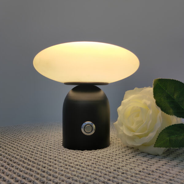 Lampe champignon avec le corps noir et le chapeau en forme d'ovale posé à l'horizontal. La lampe est allumée. Elle a un bouton gris sur le devant. A sa droite, une rose blanche. Sur fond gris.