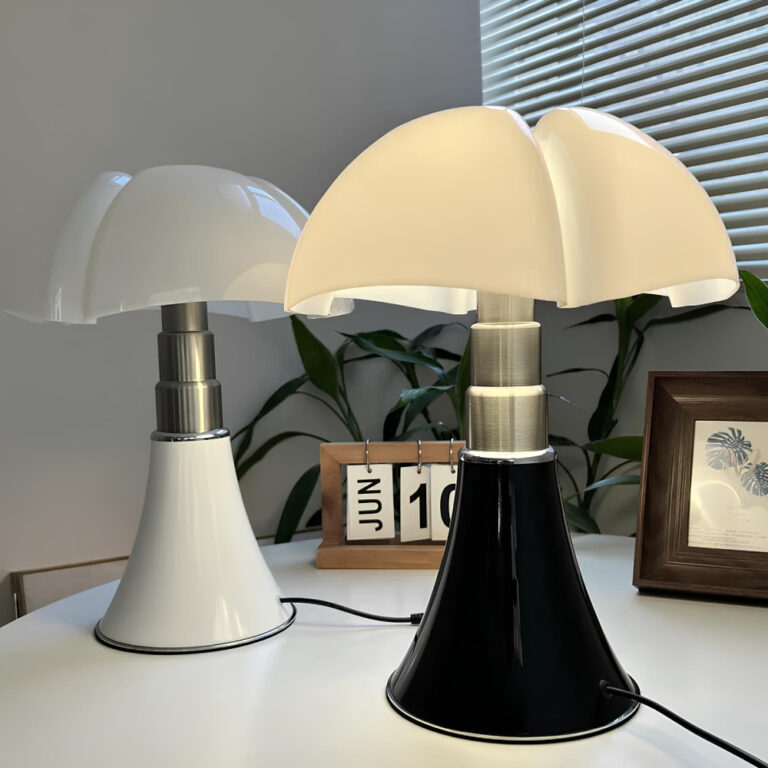 Table ronde blanche avec posées dessus deux lampes champignon au design italien. Celle de devant à droite à le corps noir et argenté avec le chapeau blanc, l'ampoule est allumée. Celle de derrière à gauche a le corps blanc et argenté et le chapeau blanc, l'ampoule est éteinte. Les deux sont reliées à leur câble noir. Derrière les lampes, un calendrier en bois avec les dates à retourner et un cadre à droite. En fond un mur blanc et une plante verte.