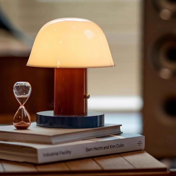Lampe champignon sans fil avec socle bleu, corps marron et chapeau blanc. L'ampoule est allumée. La lampe est posée sur deux livres superposés sur un support en bois. Un sablier à gauche. Le fond est fou.