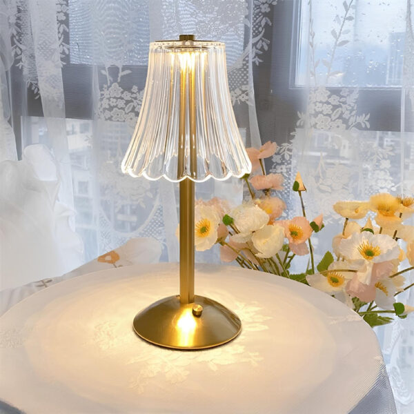 Lampe champignon vintage avec socle rond bouton et tige dorés et abat-jour transparent. Elle est allumée et posée sur une table blanche ronde devant une fenêtre avec voilages blancs avec des fleurs pastel derrière.