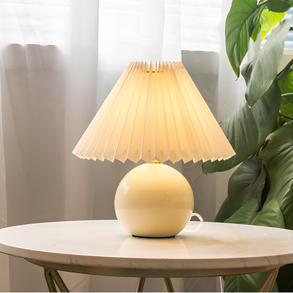 Lampe champigonn avec un corps en forme de boule en céramique beige et un abat-jour beige plissé. L'ampoule est allumée. La lampe est posée sur une table ronde couleur bois clair. Derrière un voilage blanc et à droite une plante verte.