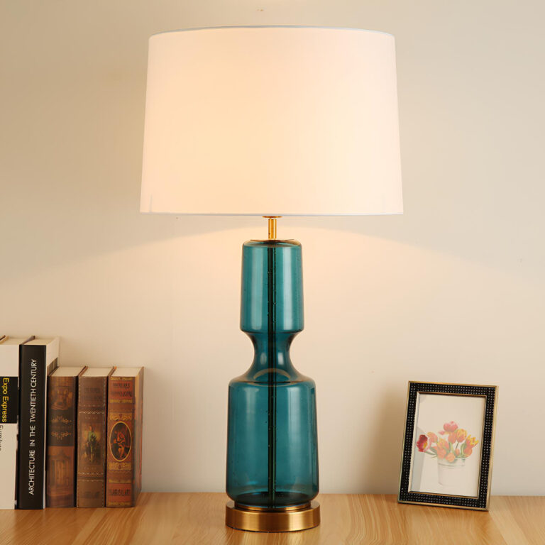 Lampe champignon vintage avec pied en verre de couleur turquoise en forme de vase translucide. Par-dessus, un abat-jour blanc en tissu satiné. La lampe est posée sur un meuble couleur bois. A sa droite, un petit cadre, à sa gauche des livres anciens. Le mur derrière est blanc.