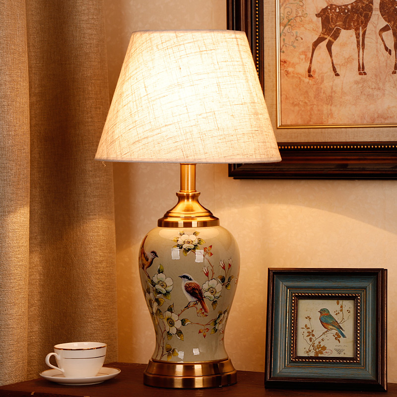 Lampe de bureau vintage avec un corps en céramique au design inspiré de motifs asiatiques tels que des fleurs de cerisier et des oiseaux sur fond beige. Son abat-jour en forme de chapeau beige en tissu cache l'ampoule allumée diffusant la lumière. La lampe est posée sur un meuble marron foncé avec une tasse blanche à gauche et un petit cadre carré à droite. A gauche un rideau marron clair, à droite sur le mur un tableau avec un encadrement marron foncé. Le mur est beige.