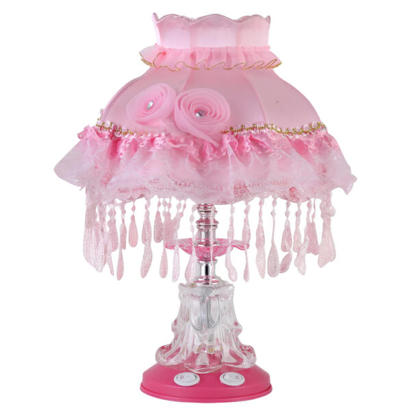 Lampe champignon de couleur rose pâle avec un socle rond rose muni de 2 boutons. Le pied en plastique transparent est surmonté d'un abat-jour en dentelle rose avec des pampilles tout le tour. Sur fond blanc.