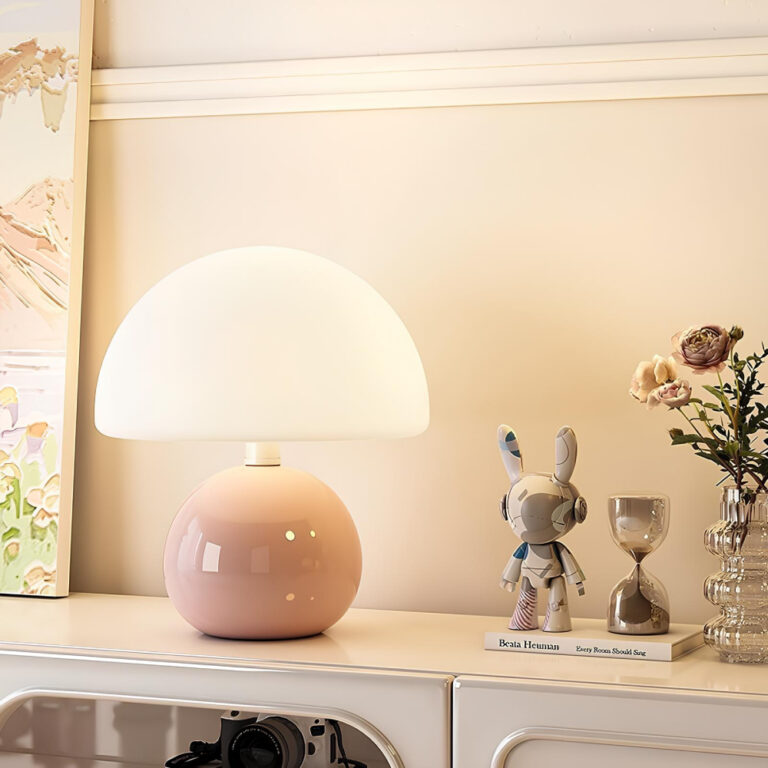 Lampe champignon avec corps rond de couleur rose pastel en céramique. Son chapeau diffuse une lumière blanc chaud. Elle est posée sur un meuble beige avec 2 figurines à sa droite et une fleur dans un récipient. A gauche un tableau. Le mur derrière est beige.