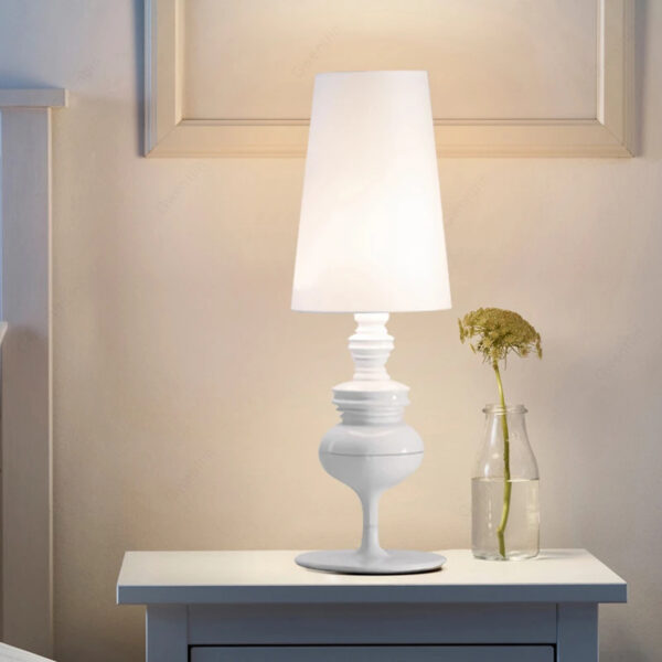Lampe de table champignon de couleur blanche avec un design élancé style vintage et un abat-jour en tissu blanc. L'ampoule est allumée. La lampe est posée sur une table de chevet blanche avec une petite fleur à droite dans un récipient transparent. Le mur derrière est blanc.