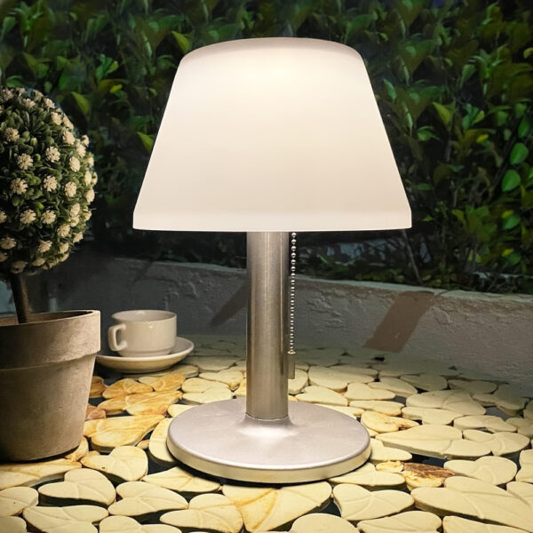 Lampe de table champignon avec socle rond et pied gris. L'abat-jour diffuse la lumière. La lampe sans fil est posée sur une table ronde avec des motifs de coeurs beige sur fond noir. A gauche une plante verte et une tasse blanche. En fond un buisson.