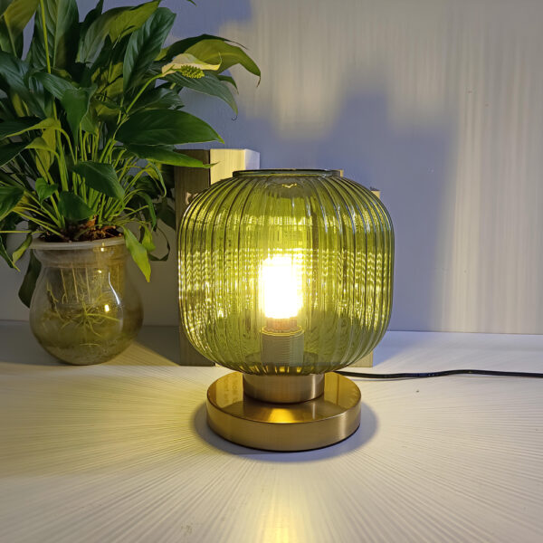 Lampe champignon avec socle rond doré surmonté d'un verre vert en forme de boule du style carré arrondi avec des stries verticales. L'ampoule est allumée. La lampe est posée sur un support blanc, au fond à gauche une plante verte dans un pot en céramique beige.