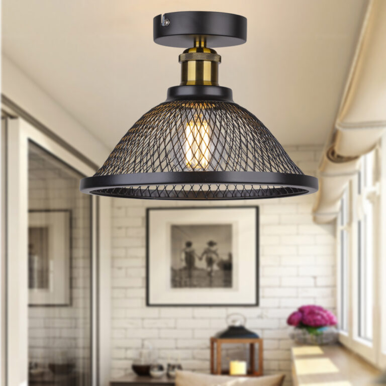 Plafonnier champignon au design industriel en métal noir, avec abat-jour en forme de grillage. Une ampoule allumée style Edison. Le plafonnier est fixé dans une véranda avec une baie vitrée sur la gauche et des fenêtres sur la droite. Au fond un mur en pierre claire et un tableau au centre. En dessous, une table basse avec des objets. A droite sur le rebord de la fenêtre une plante fleurie violette.